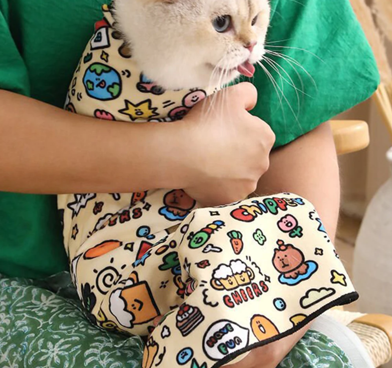 Cat wearing a colorful Magic Pet Wrap with cartoon designs, held by a person in a green shirt.